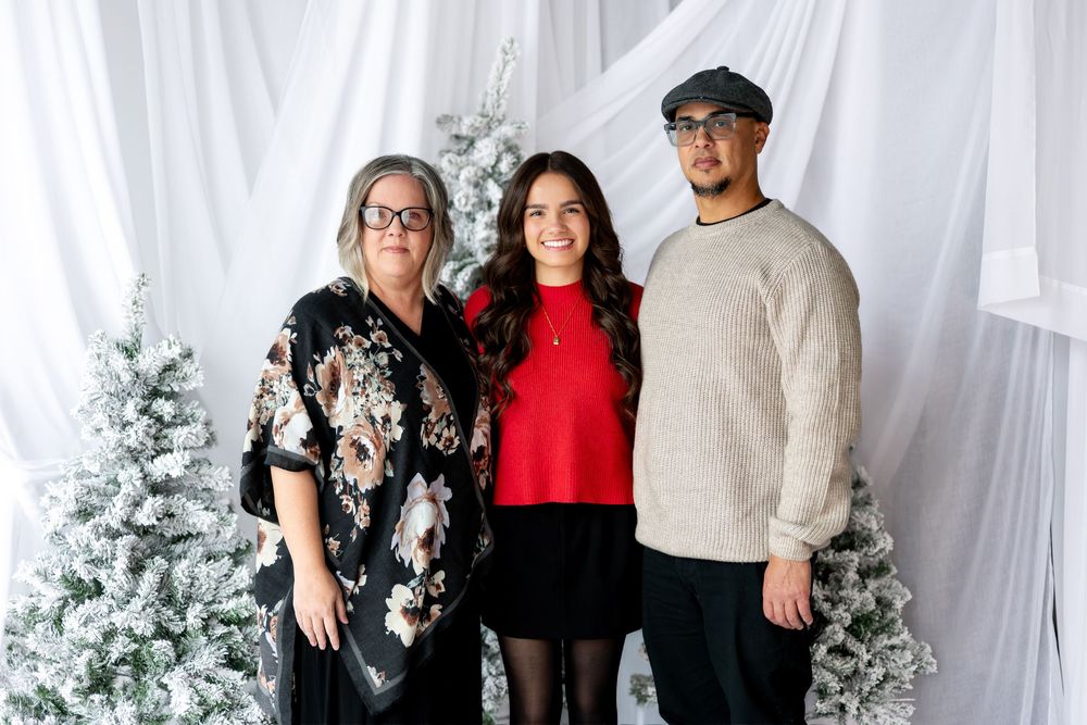 Family of three poses in holiday outfits before snow-flocked Christmas trees in studio setting.