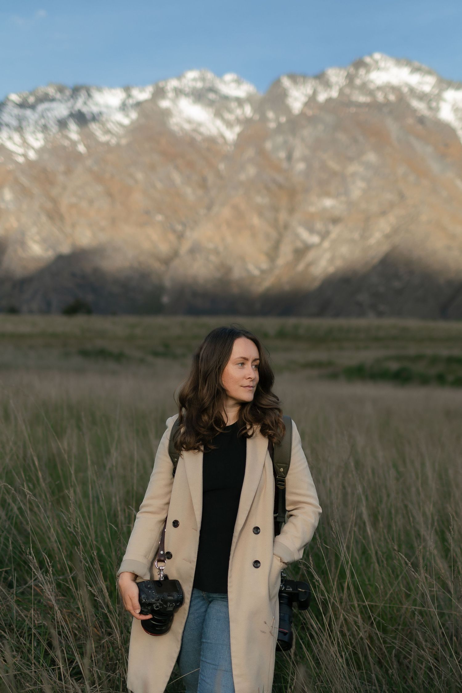 A photographer stands in a grassy field with snow-capped mountains in the background wearing a beige coat and black top.