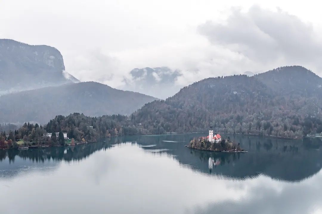 Lake Bled church with mountains behind, reflected in calm lake water.
