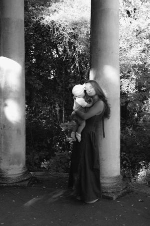 Black and white photo of a mother holding and kissing her baby between stone columns in a garden.