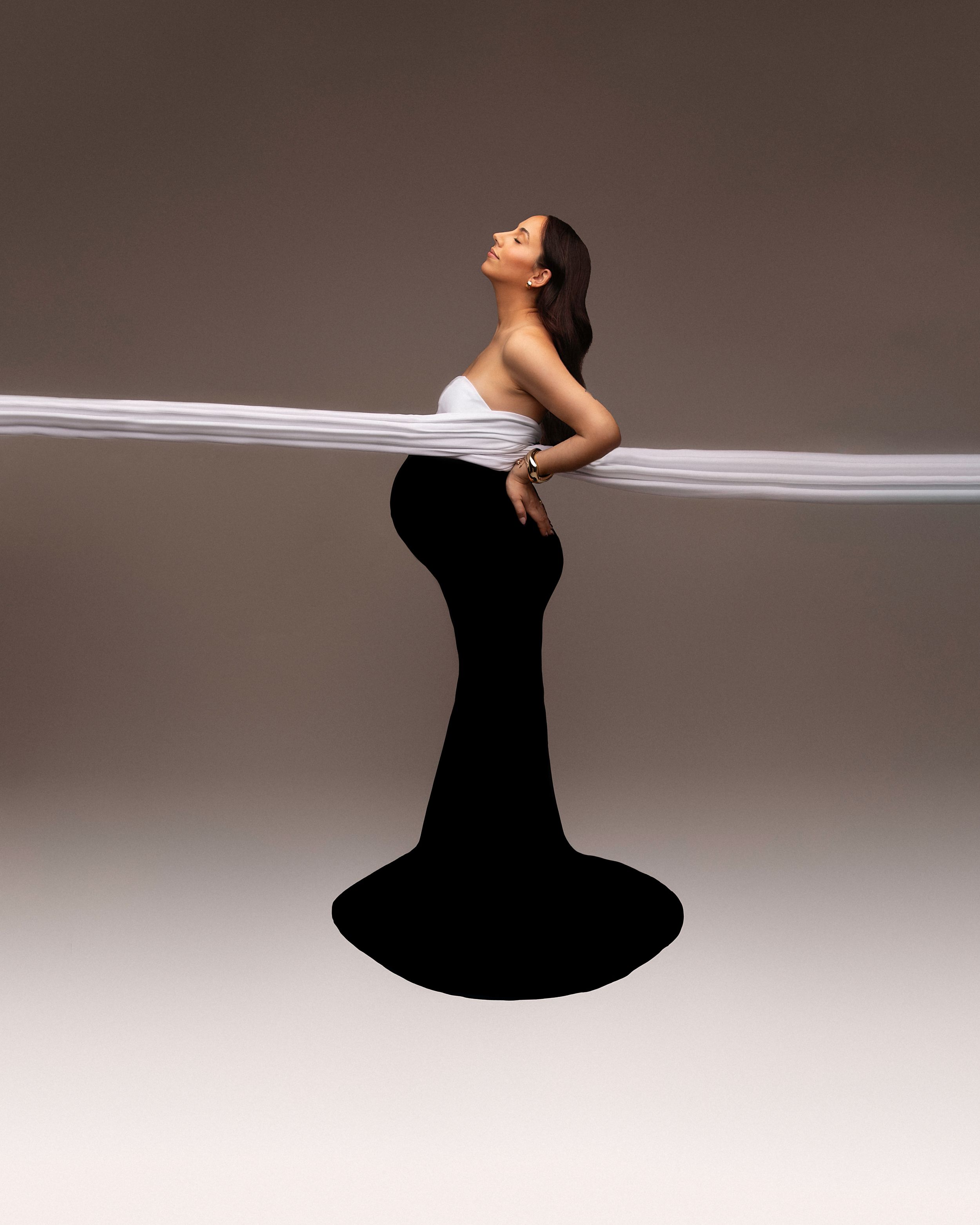 Pregnant woman in black mermaid skirt with white fabric streaming across a studio background.