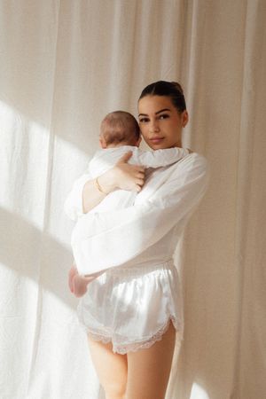 Young mother in white shirt and satin shorts holding newborn baby near sheer curtains in soft light.