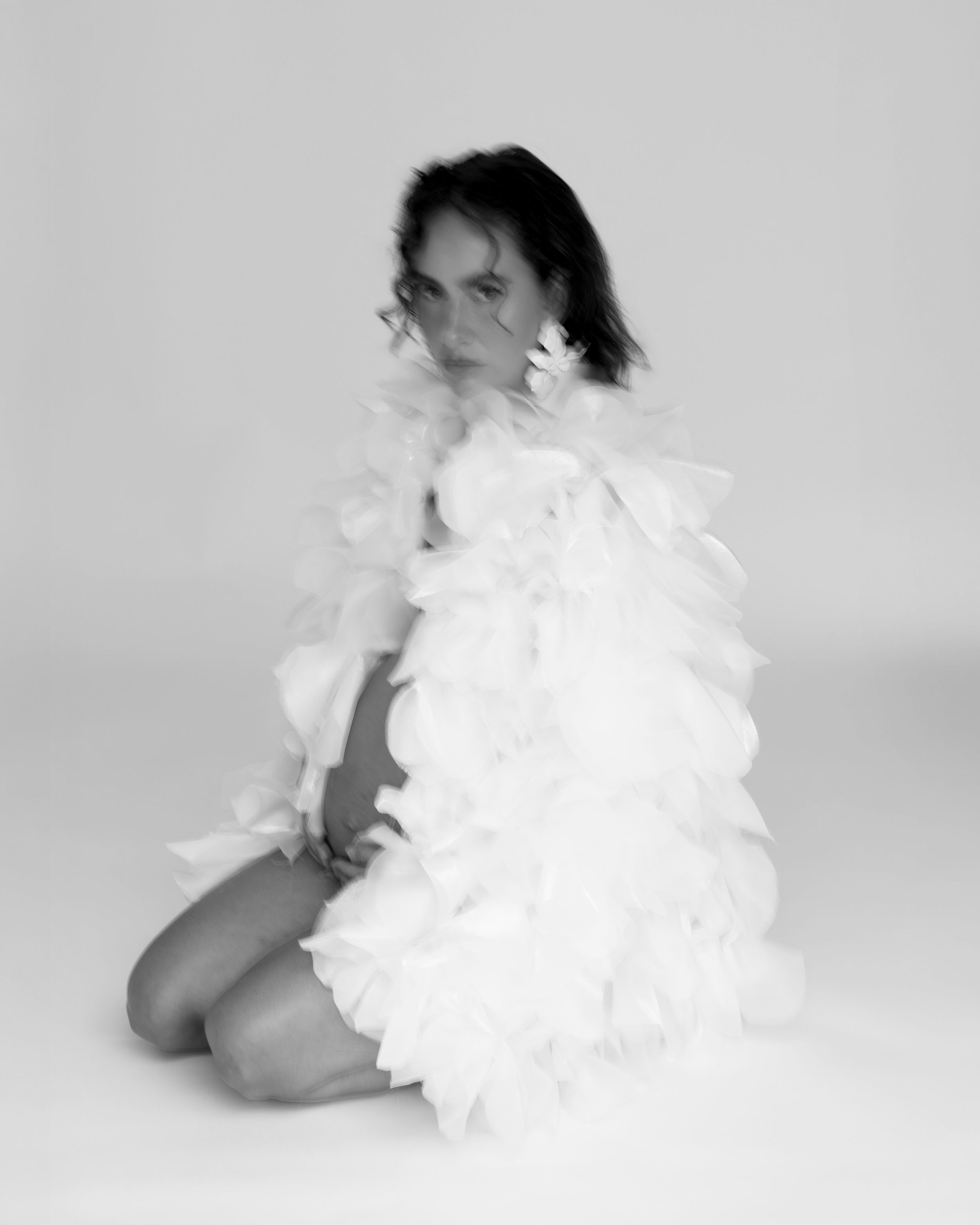 Black and white photo of a woman sitting gracefully in a voluminous white ruffled feather dress against a light background.