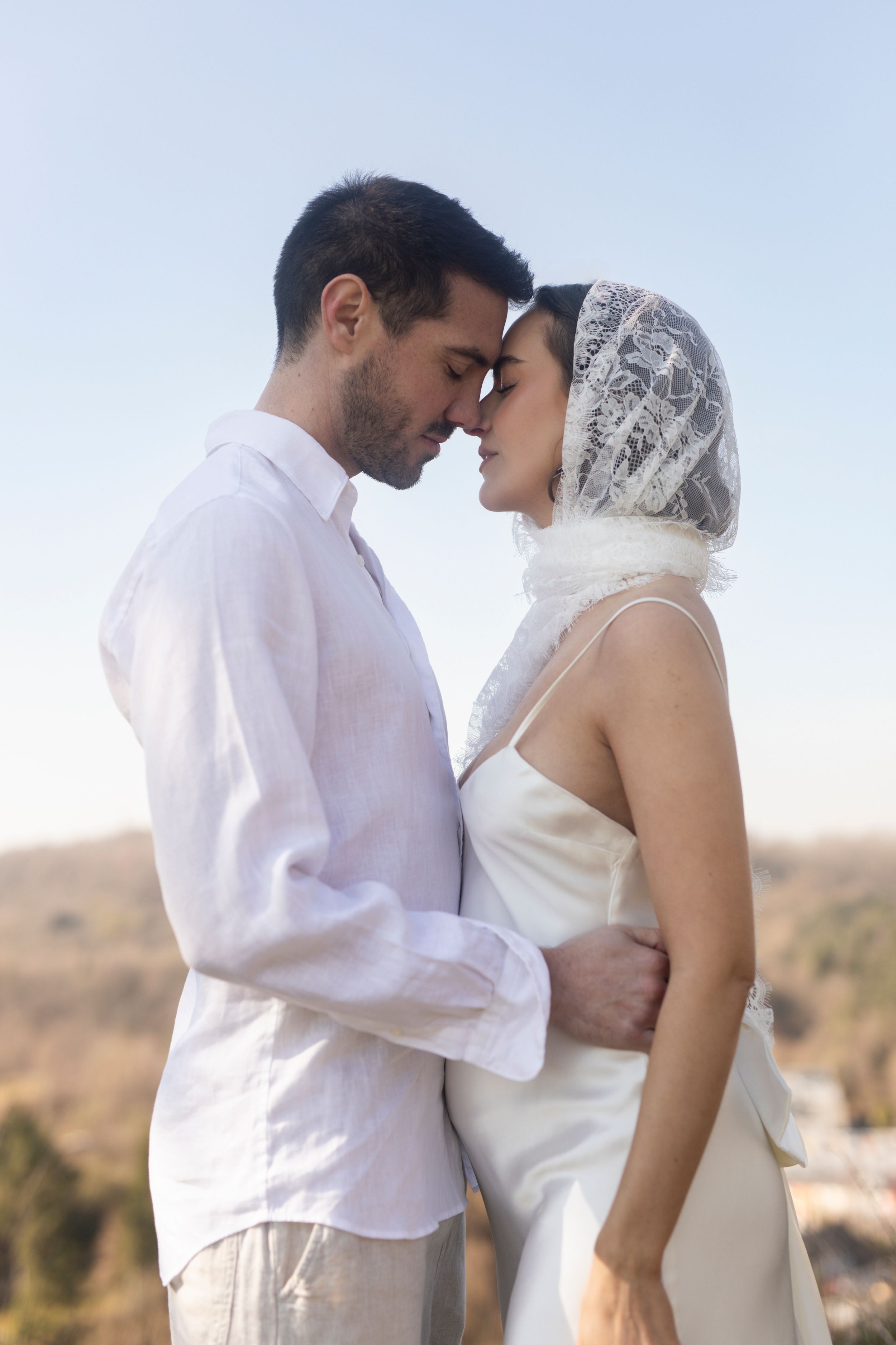 Bride and groom in white attire sharing romantic forehead touch outdoors under clear blue sky.
