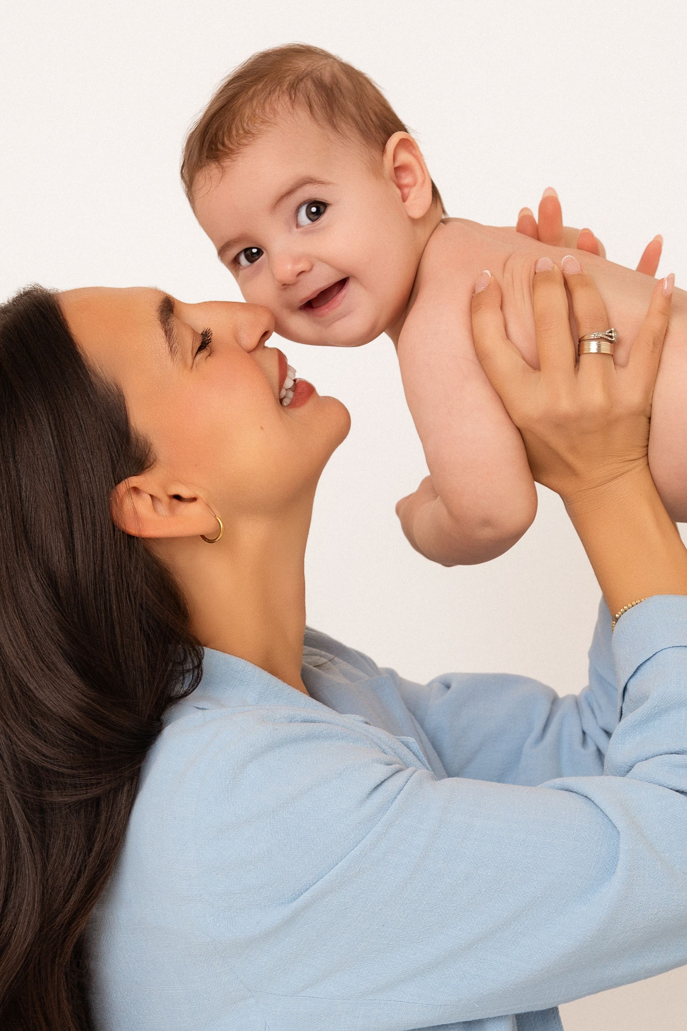 Happy mother in blue shirt lifting smiling baby up close to her face against white background.