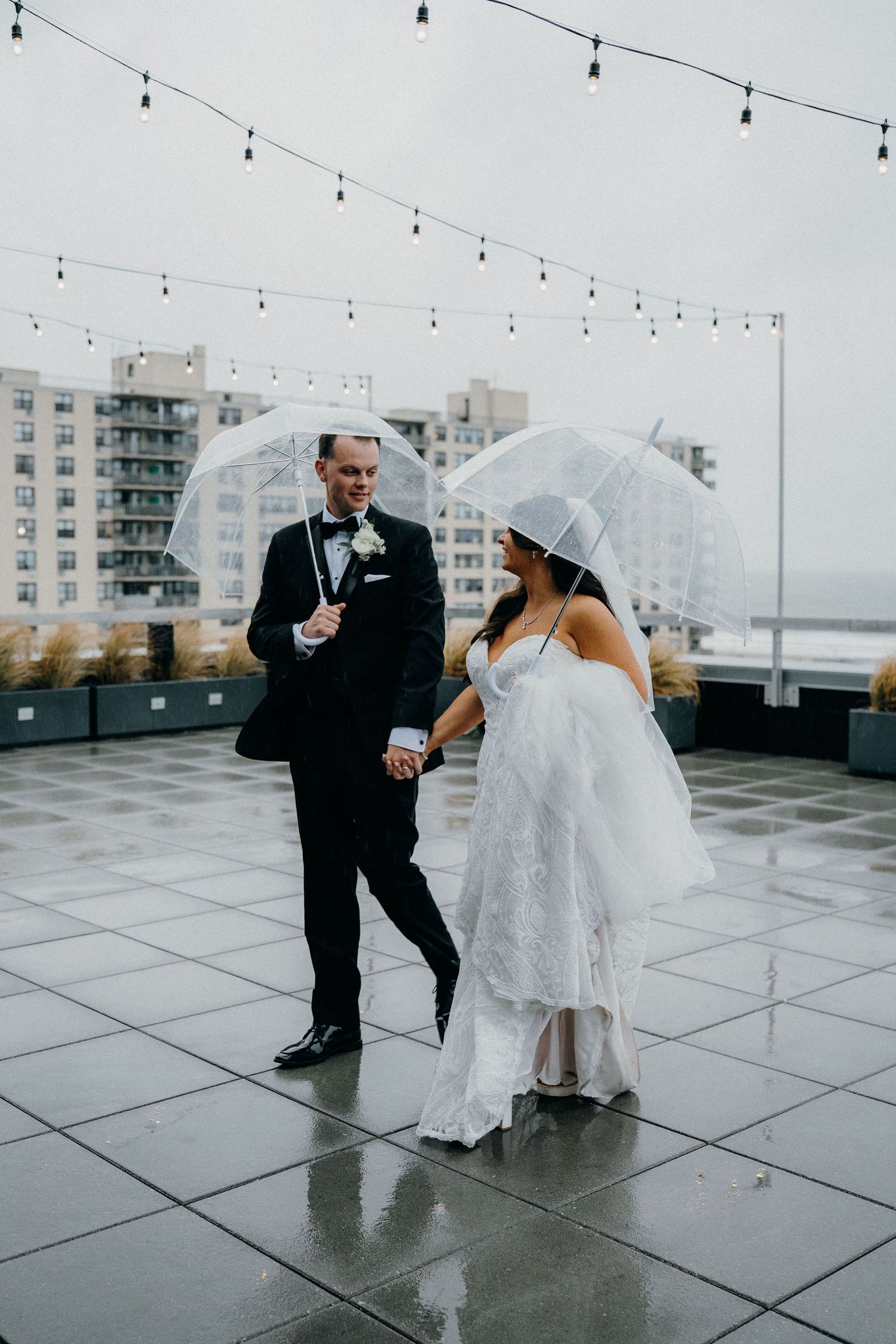 A couple in wedding attire dances playfully in the rain on a rooftop terrace with string lights and city buildings visible.