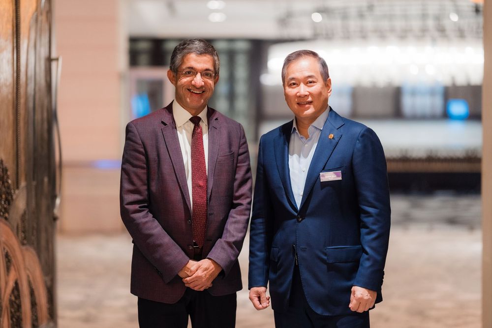 Two professionals in business attire shake hands in a corporate office hallway setting.