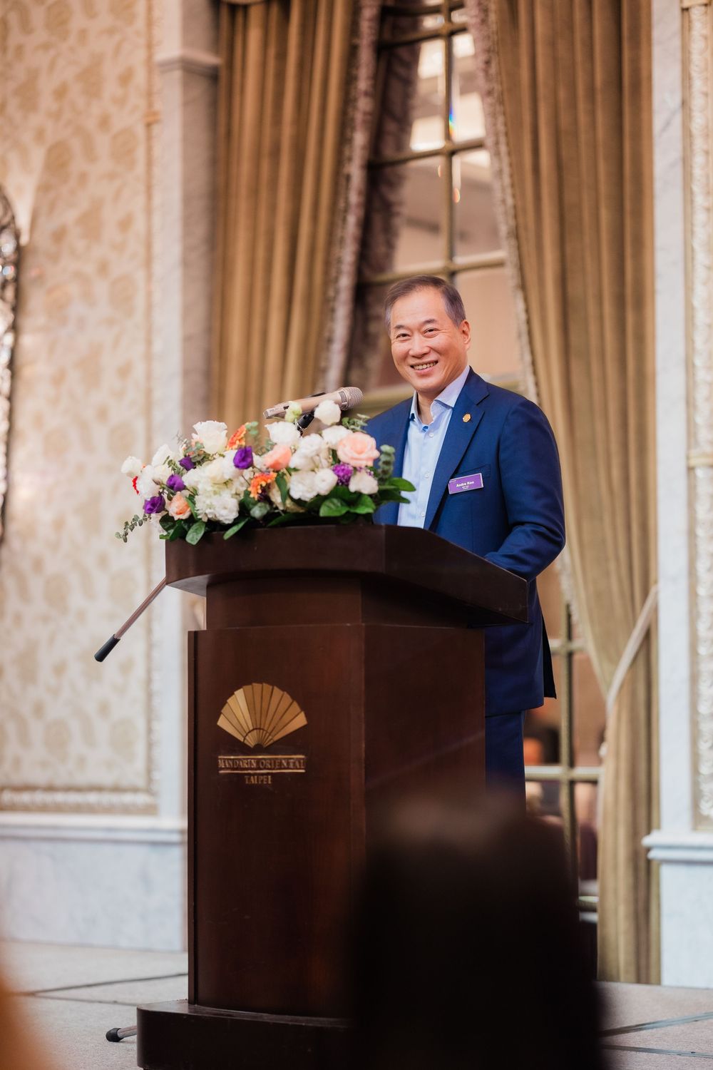 A speaker stands at a wooden podium decorated with floral arrangements in an elegant room with patterned wallpaper.
