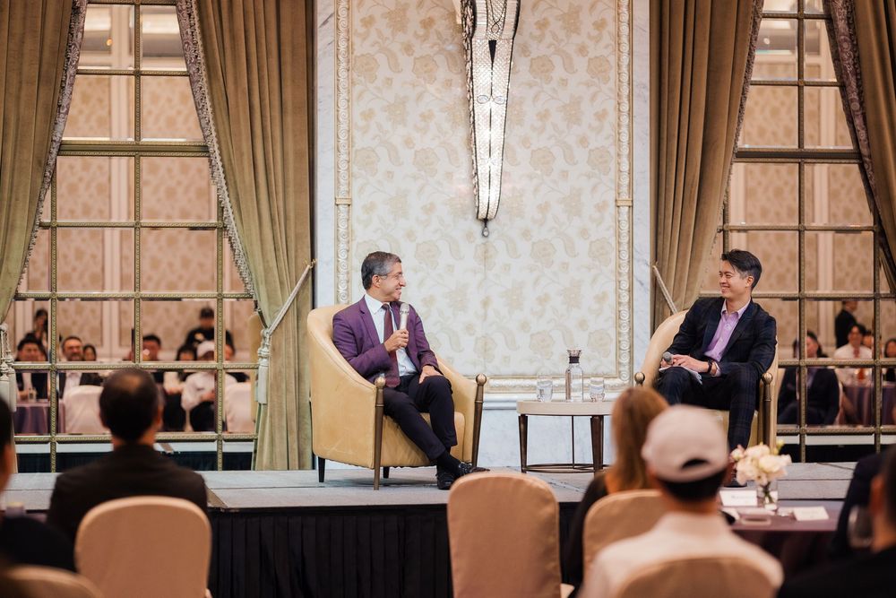 Professional speaker presenting to an audience in an elegant conference room with ornate wallpaper and drapes.