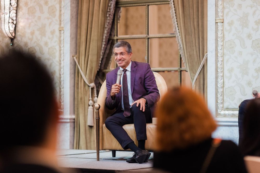 Speaker gesturing while giving presentation in an upscale venue with decorative wall panels.