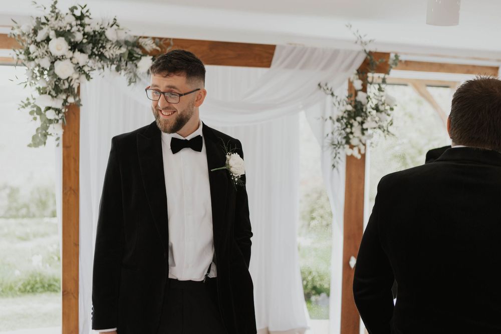 Groom in black tuxedo with bow tie and white boutonniere smiling at altar during wedding ceremony.