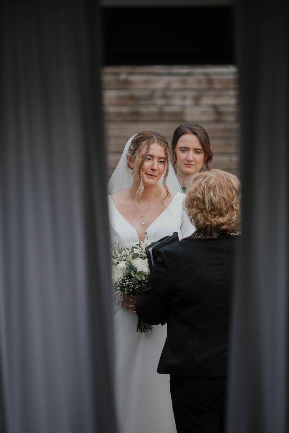 Bride in white gown and veil holding bouquet stands before officiant during intimate wedding ceremony.