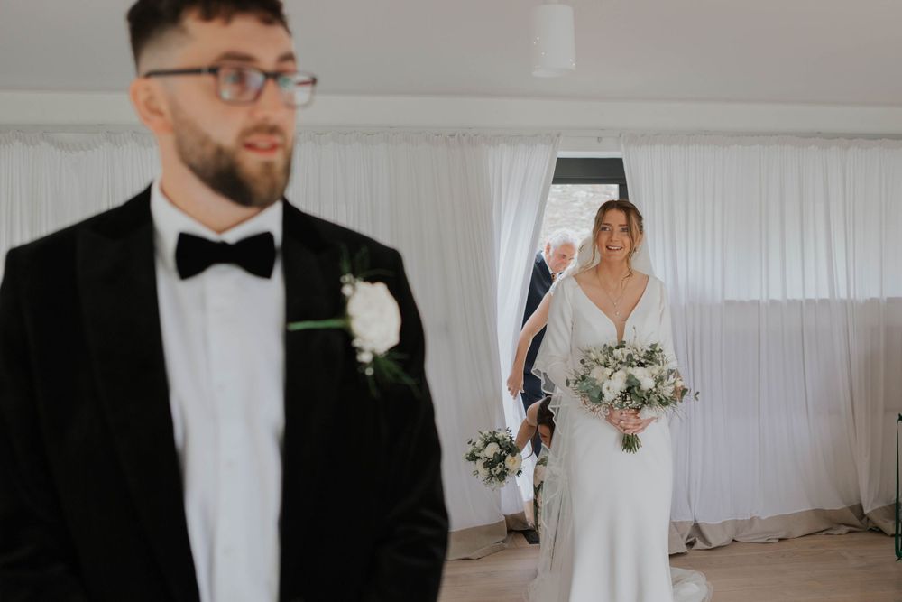Groom in black tuxedo turns as bride in white wedding dress approaches during ceremony.