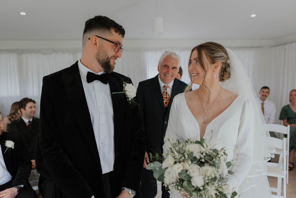 Smiling bride and groom exchange vows at altar with wedding guests seated in white chairs behind them.