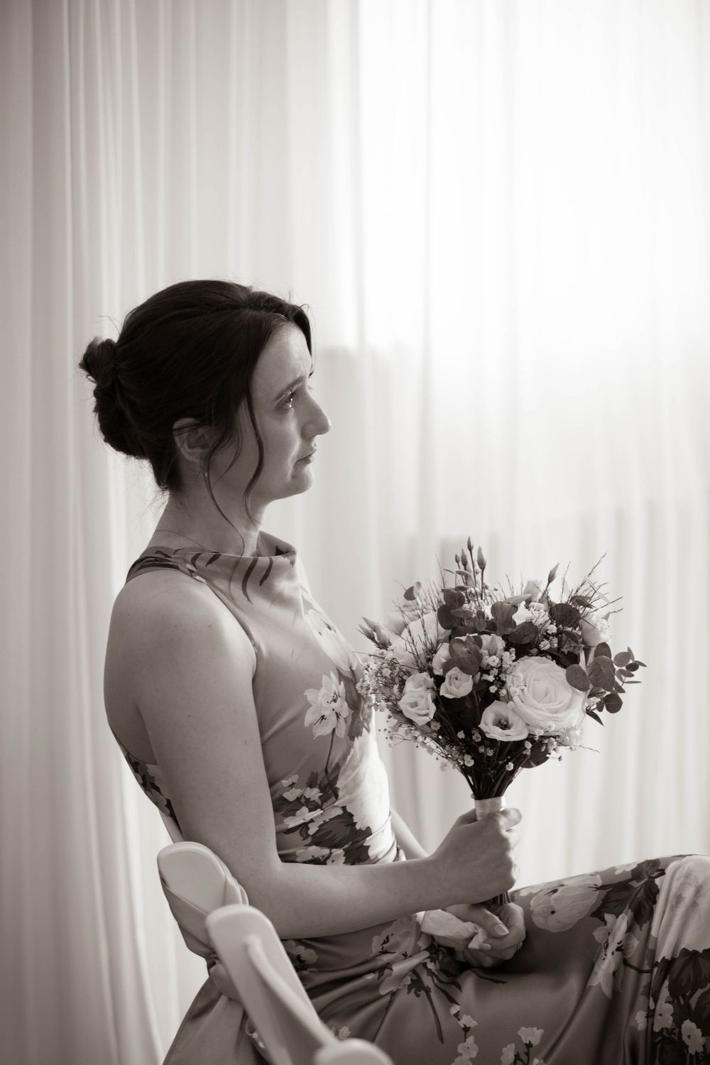 Black and white portrait of bridesmaid in floral dress seated holding a bouquet by a bright window.