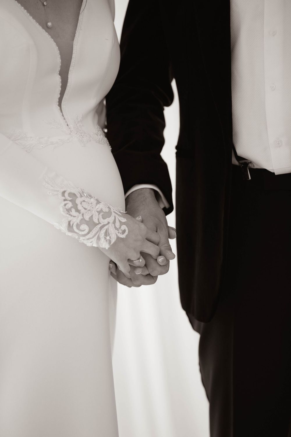 Black and white close-up of bride and groom holding hands showing lace wedding dress sleeve detail.