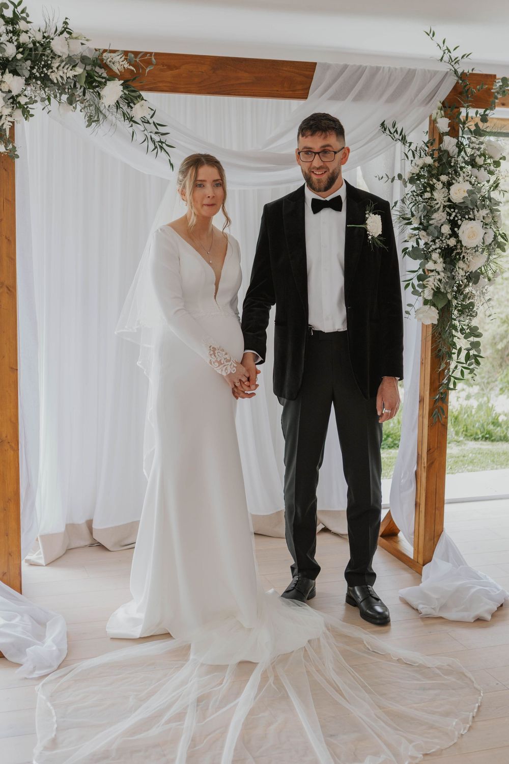 Bride and groom holding hands at wedding altar with white draping and greenery floral arch.