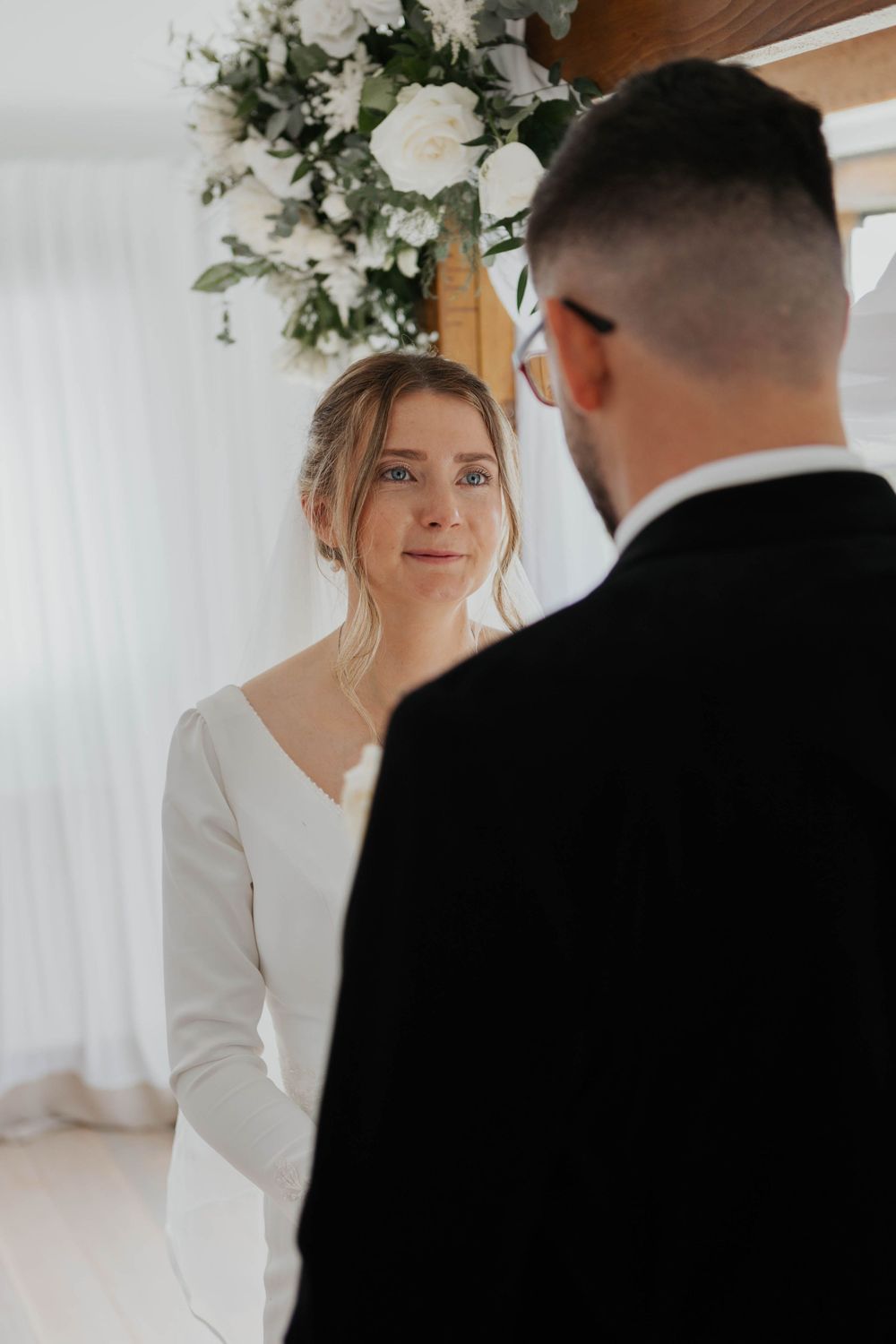 Bride in white long-sleeve gown gazing lovingly at groom during indoor wedding ceremony.