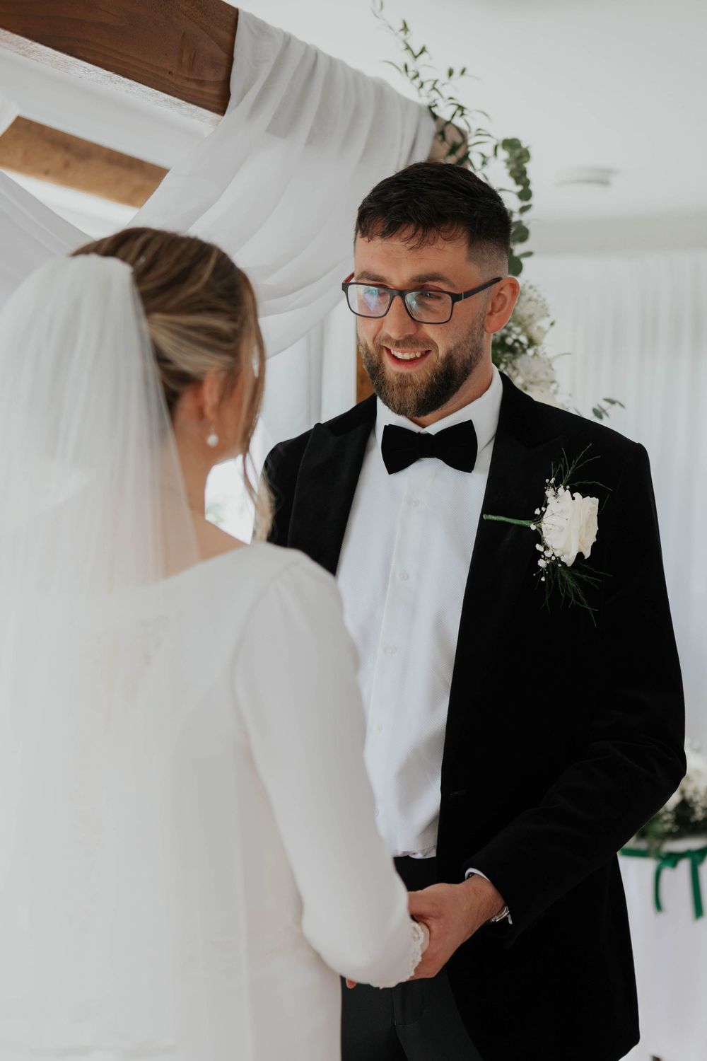 Smiling groom in black tuxedo with bow tie holding bride's hands at wedding ceremony.