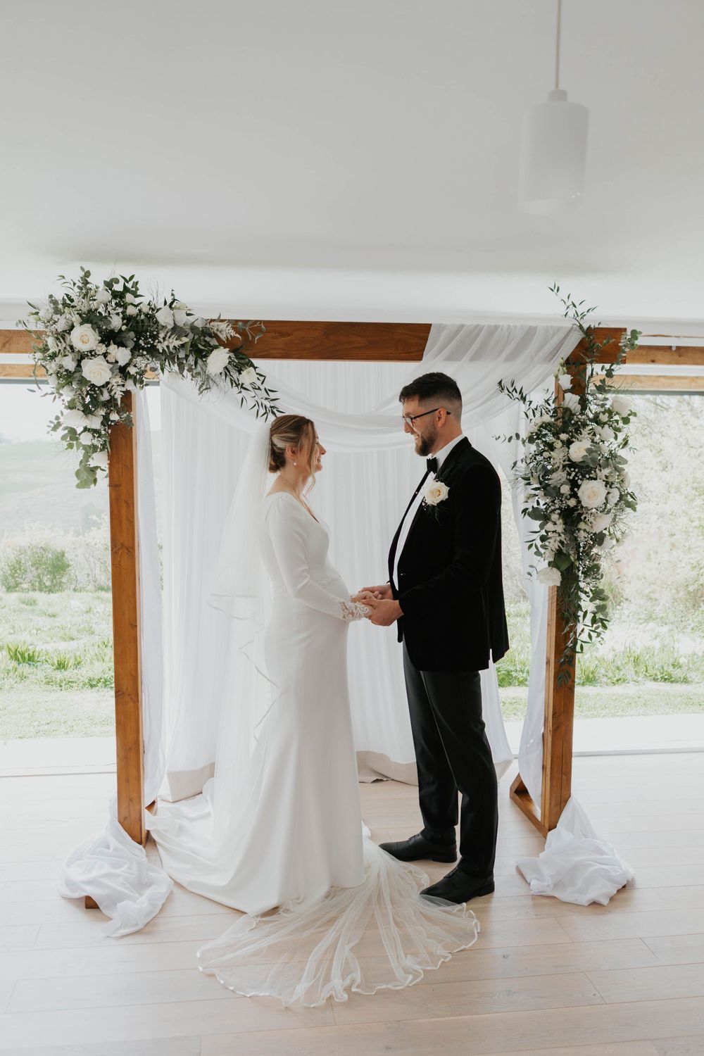 Bride and groom exchanging vows at wooden arch adorned with white roses and greenery.