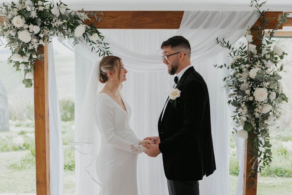 Couple holding hands at elegant wedding ceremony beneath floral arch with white roses.