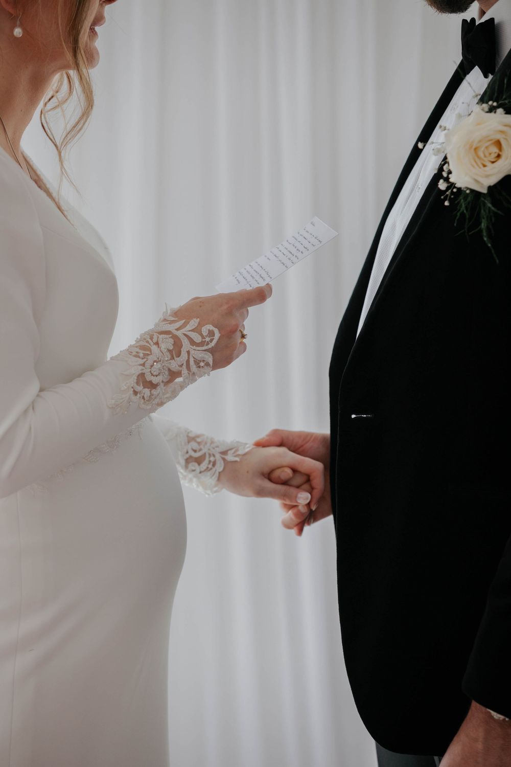 Bride in lace-sleeve wedding gown reading vows while holding groom's hand at altar.