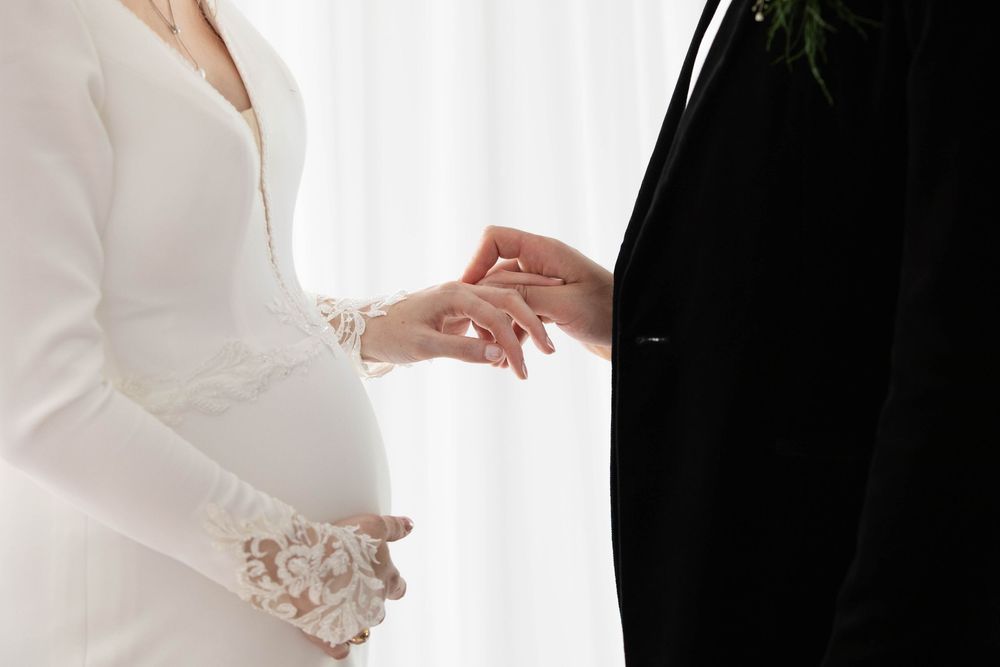 Pregnant bride placing wedding ring on groom's finger during intimate ceremony.