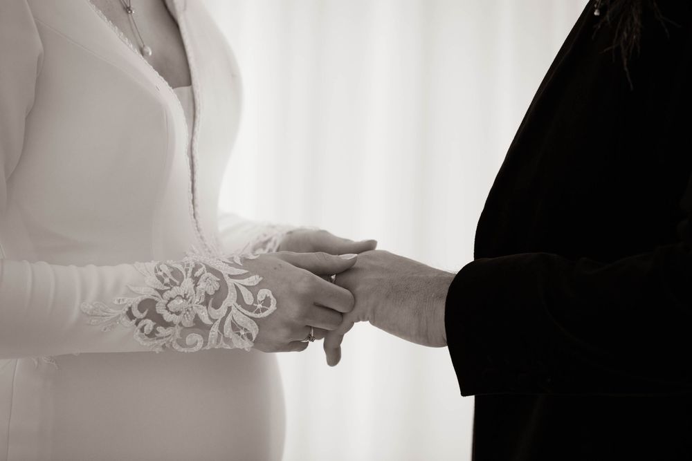 Black and white close-up of bride and groom holding hands during wedding ring exchange.