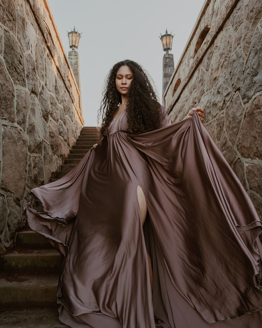 Figure in a flowing brown dress poses dramatically against stone walls and stairs in moody outdoor setting.