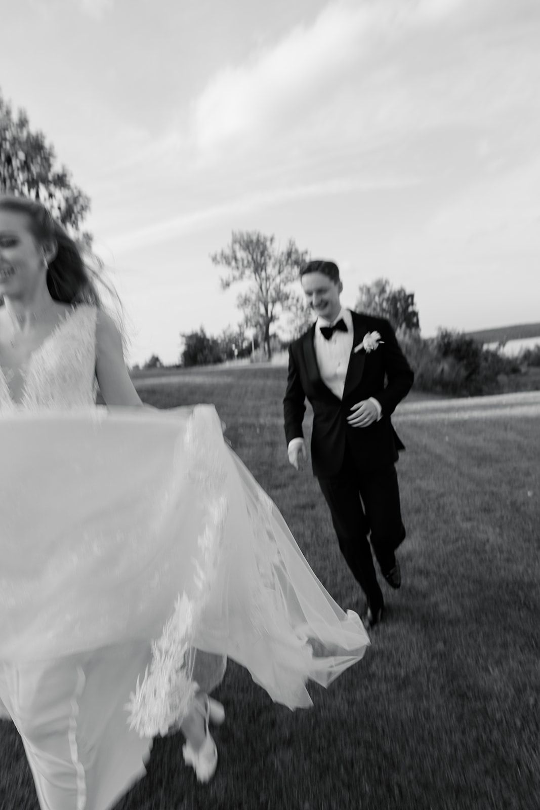 A couple in wedding attire runs playfully through a grassy field on their special day.