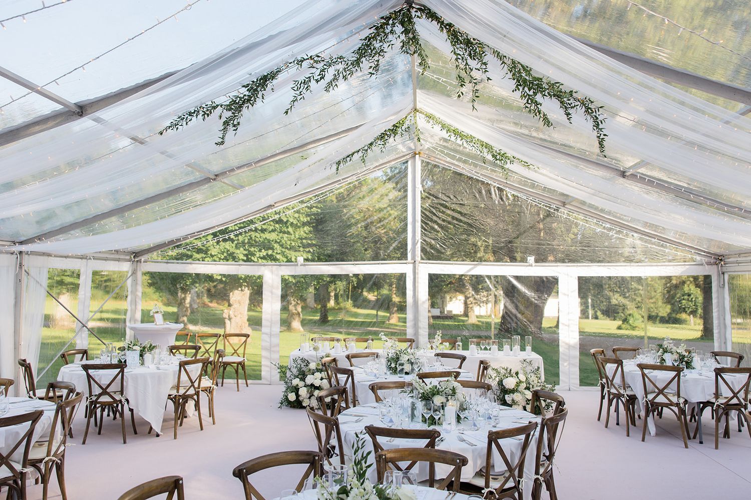 A large clear tent set up for an outdoor wedding reception with wooden cross-back chairs and round dining tables.