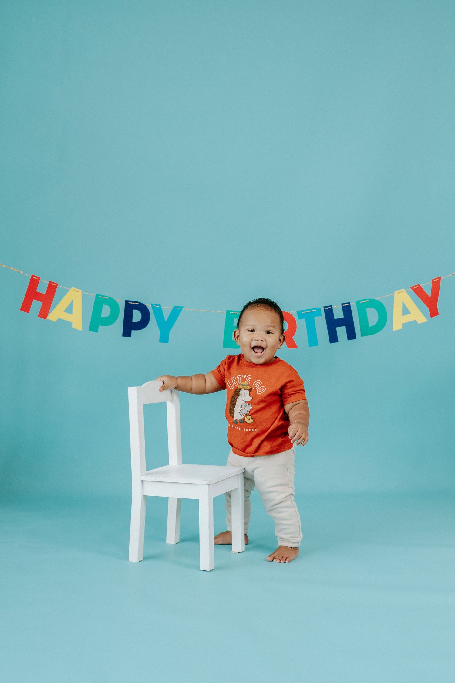 Baby in orange shirt crawls and plays near white chair with colorful happy birthday banner against turquoise background.