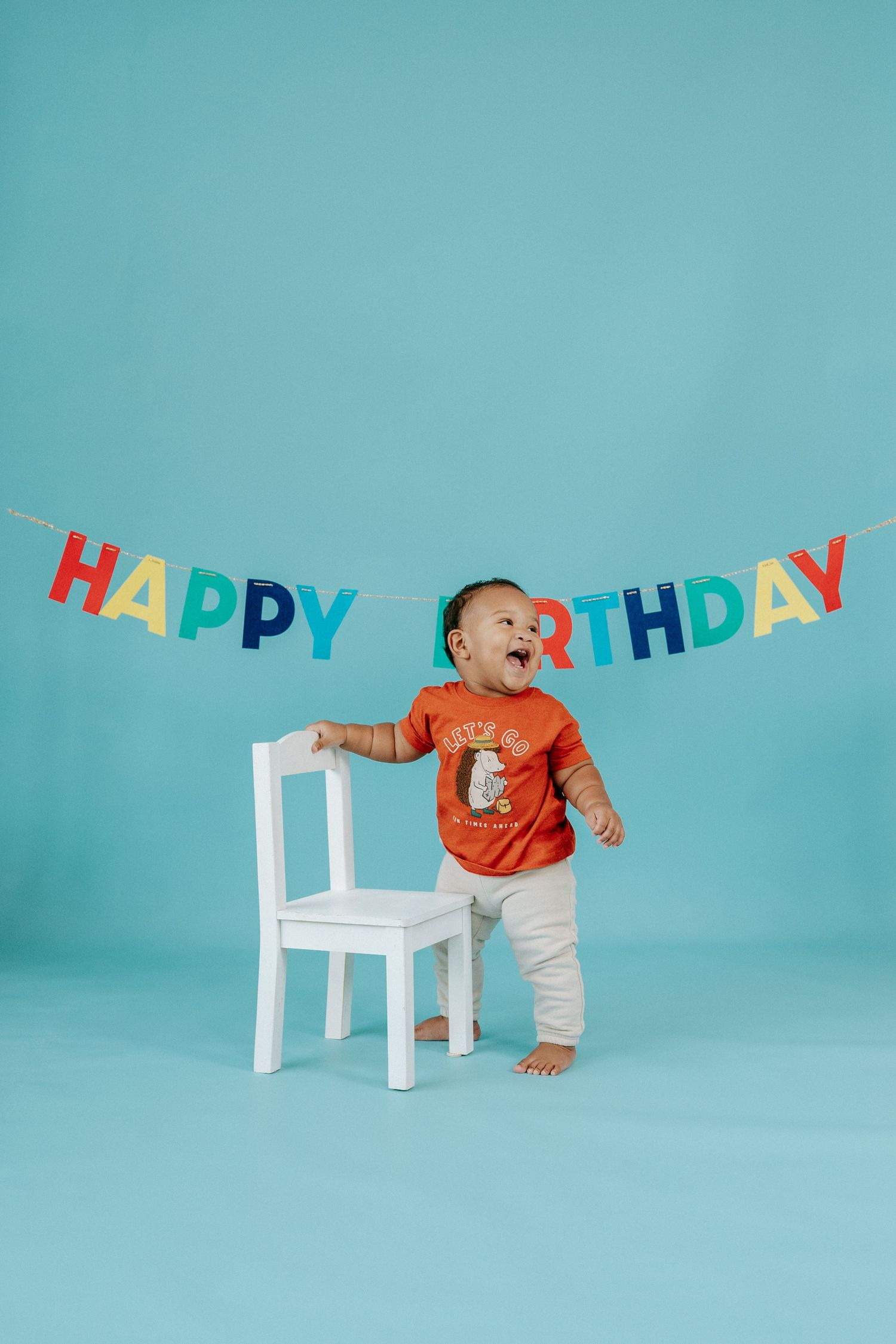 Small child in orange shirt stands beside white chair under colorful Happy Birthday banner against turquoise background.