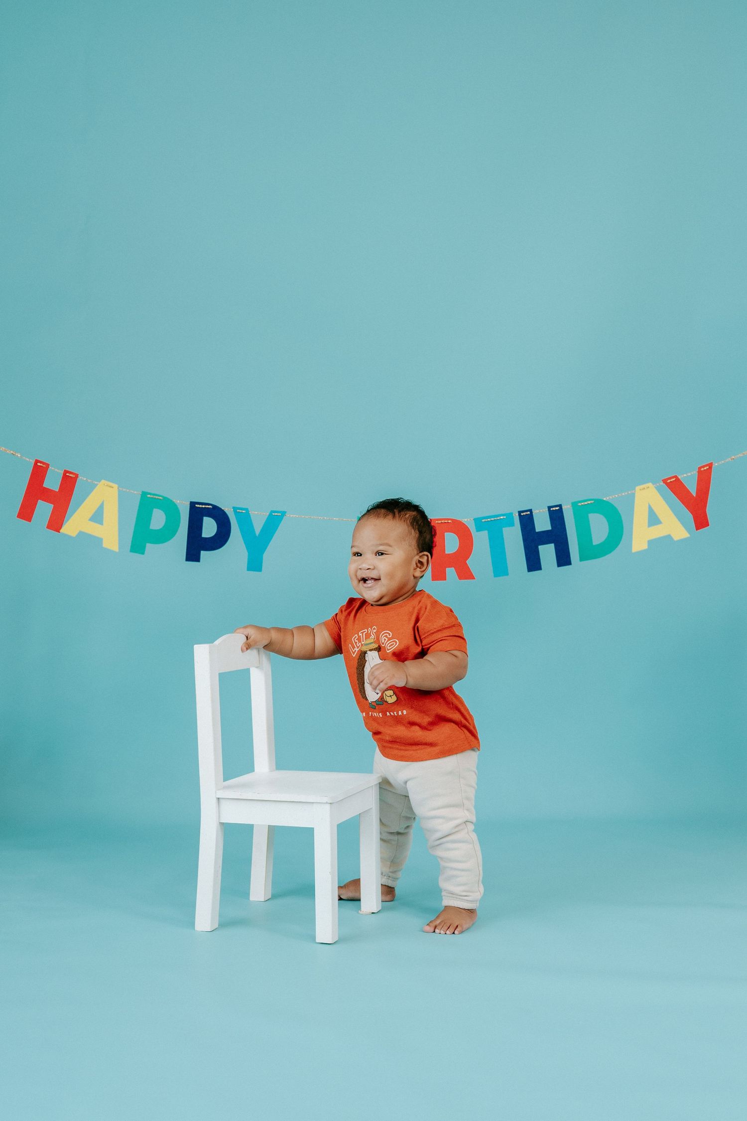 Baby in orange shirt crawls near white chair with colorful happy birthday banner on turquoise background.