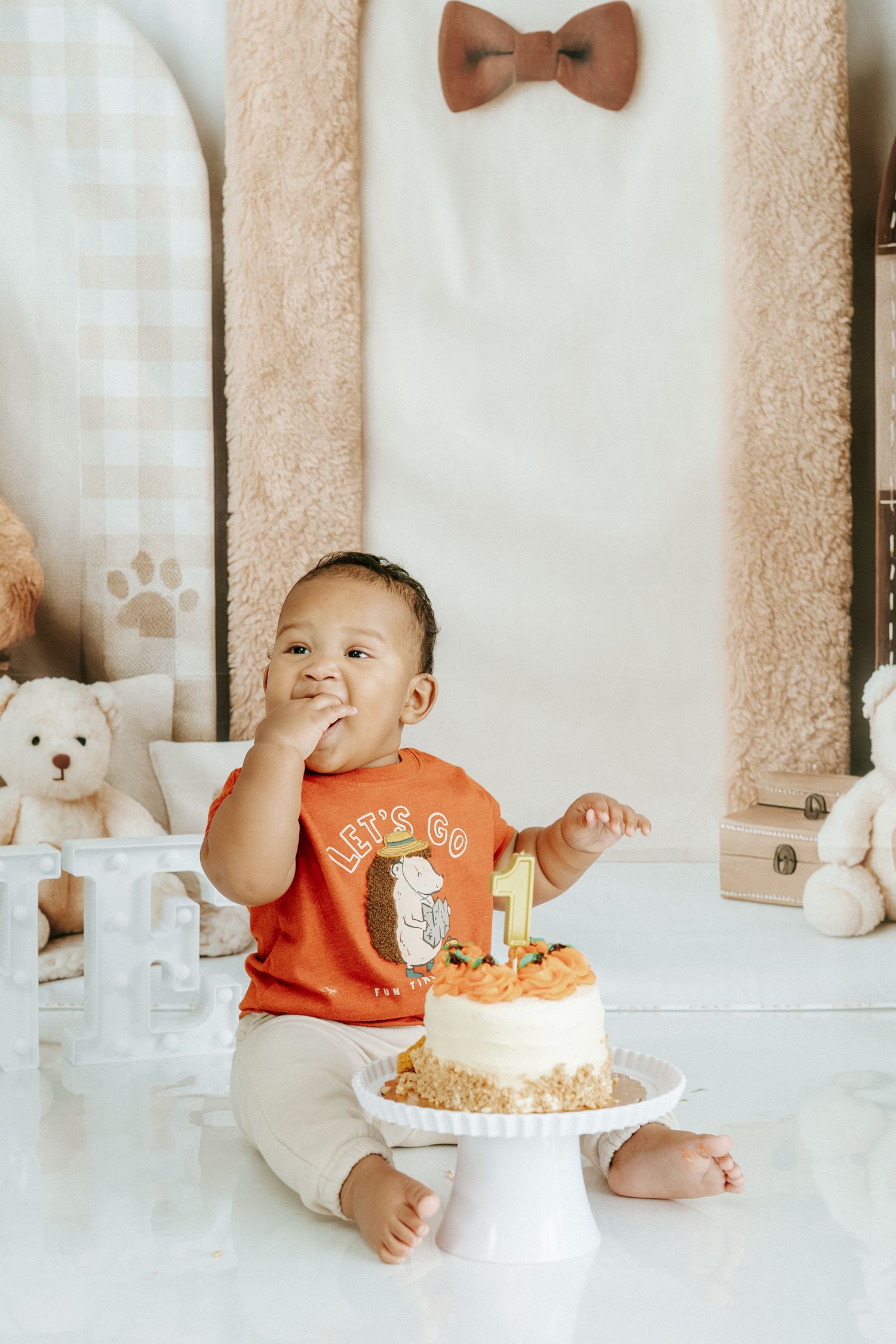 A small child in an orange shirt sits with a birthday cake on a white pedestal surrounded by stuffed animals and decor.