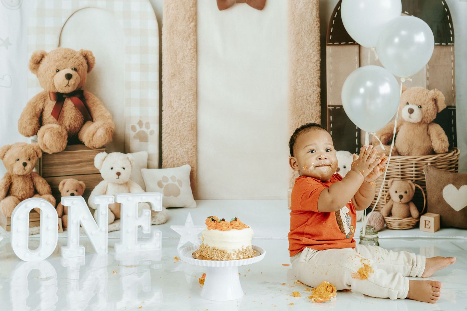 A first birthday celebration setup with teddy bears, white balloons, 'ONE' letters, and a cake on a white stand.