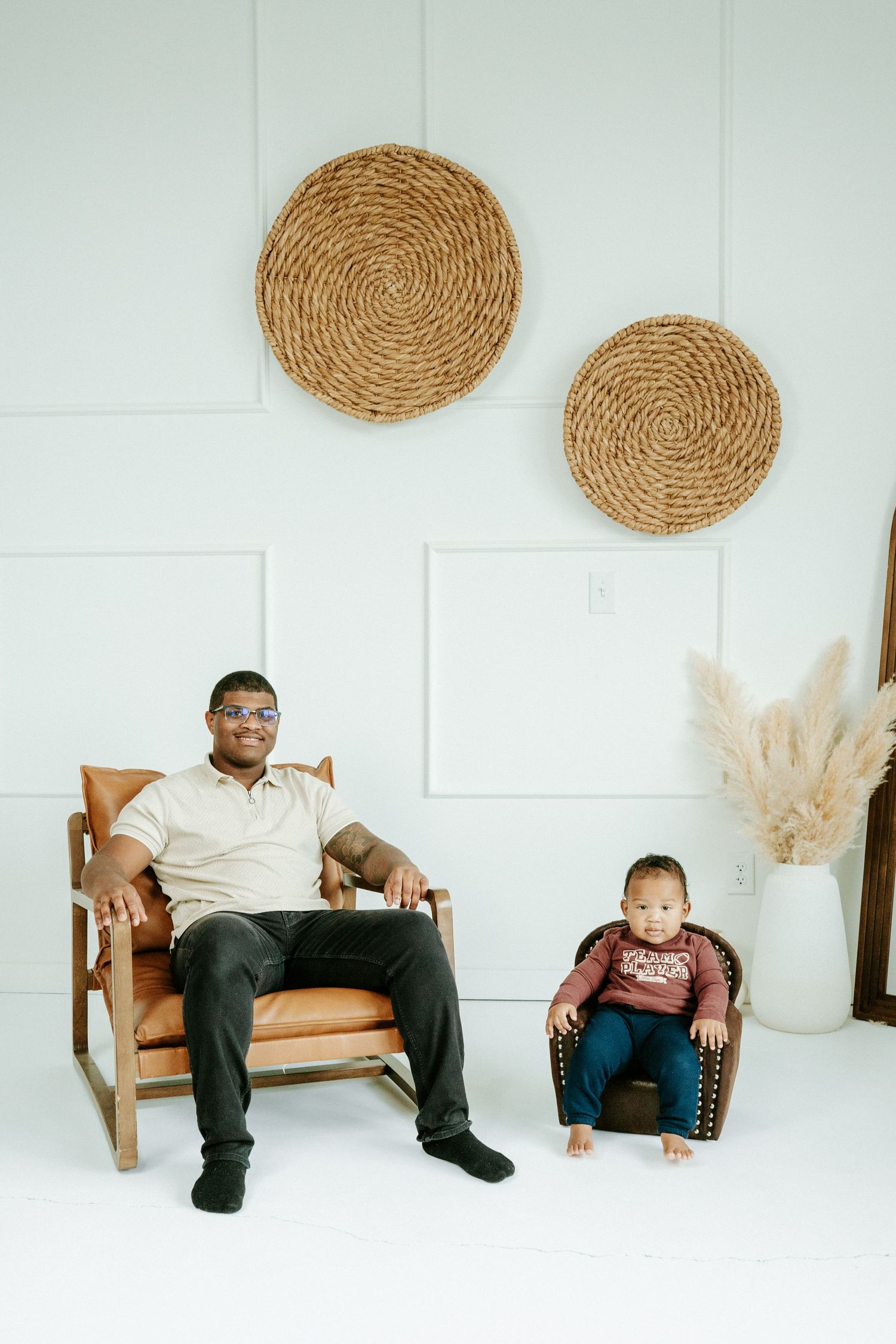 Two people sitting in matching leather armchairs against a white wall decorated with woven circular wall art.