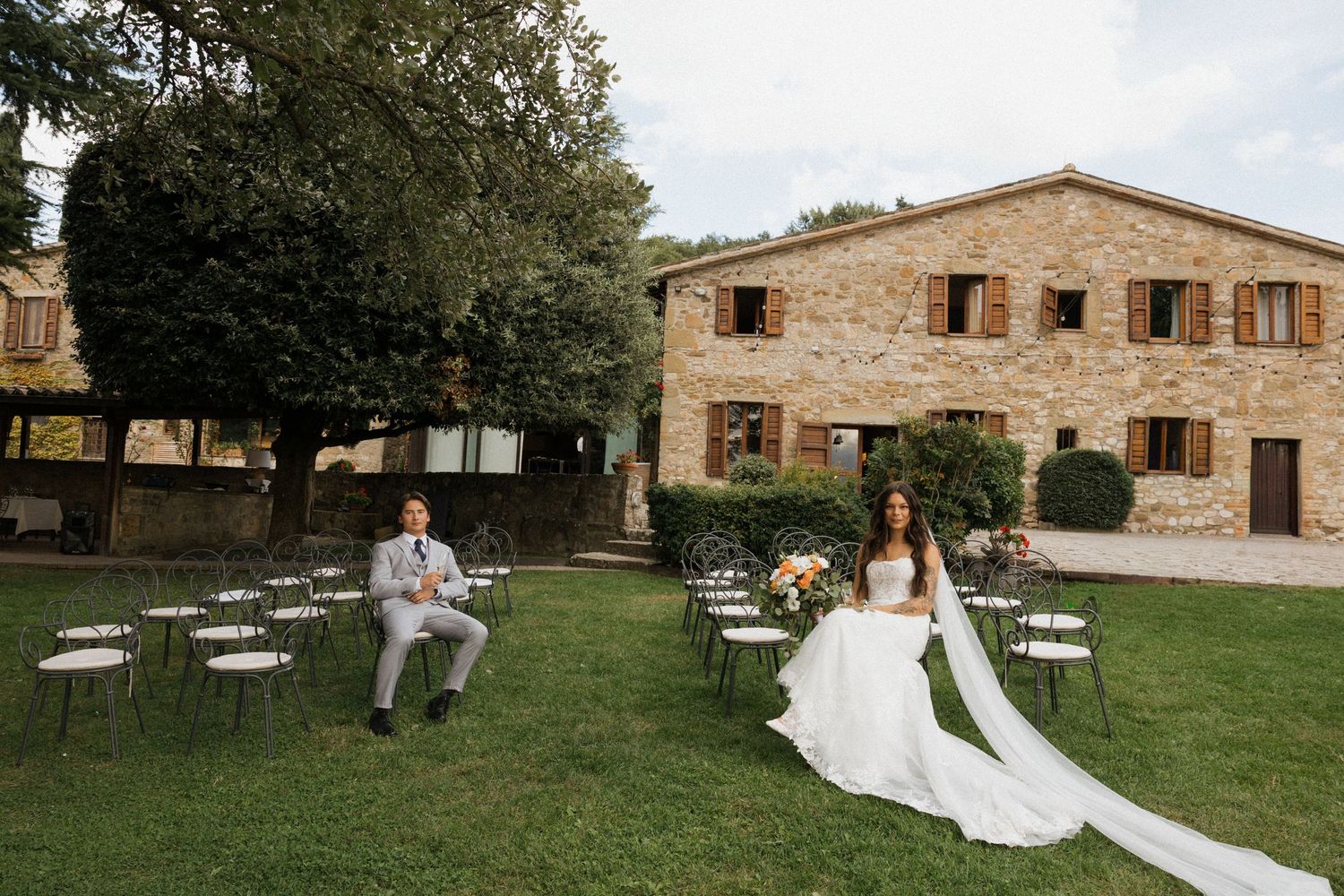 Wedding party poses in front of rustic stone building exterior with dark formal attire and yellow bouquets.