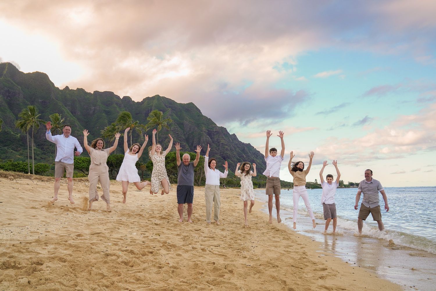 Family photo session in Kualoa regional park in white attire poses joyfully on a scenic beach at sunset with mountains in the background.