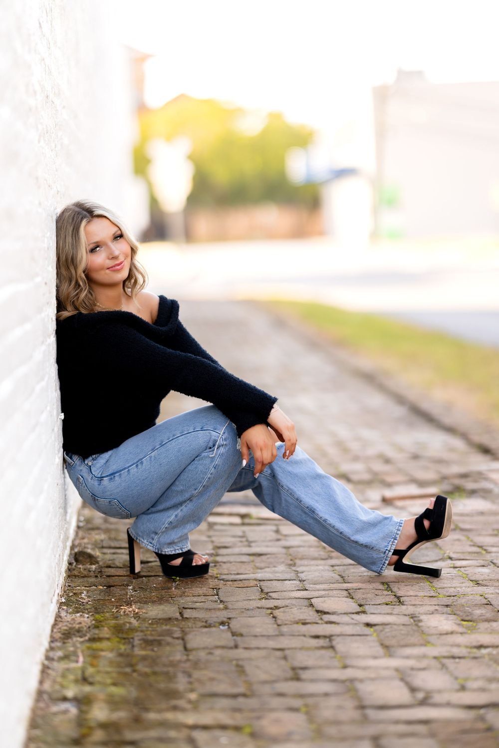 Person wearing navy top and denim sits on cobblestone path during outdoor photoshoot.