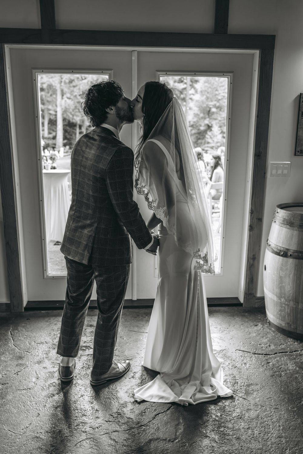 Black and white photo of a bride and groom sharing a kiss in front of French doors on their wedding day.