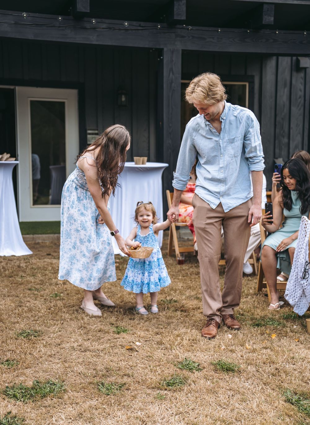 A couple in summer attire helps a toddler girl in a blue dress walk during an outdoor wedding celebration.