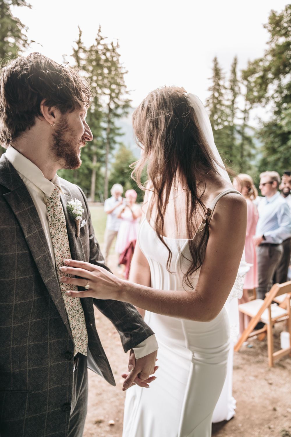 Bride and groom hold hands during outdoor wedding ceremony surrounded by tall pine trees and seated guests.