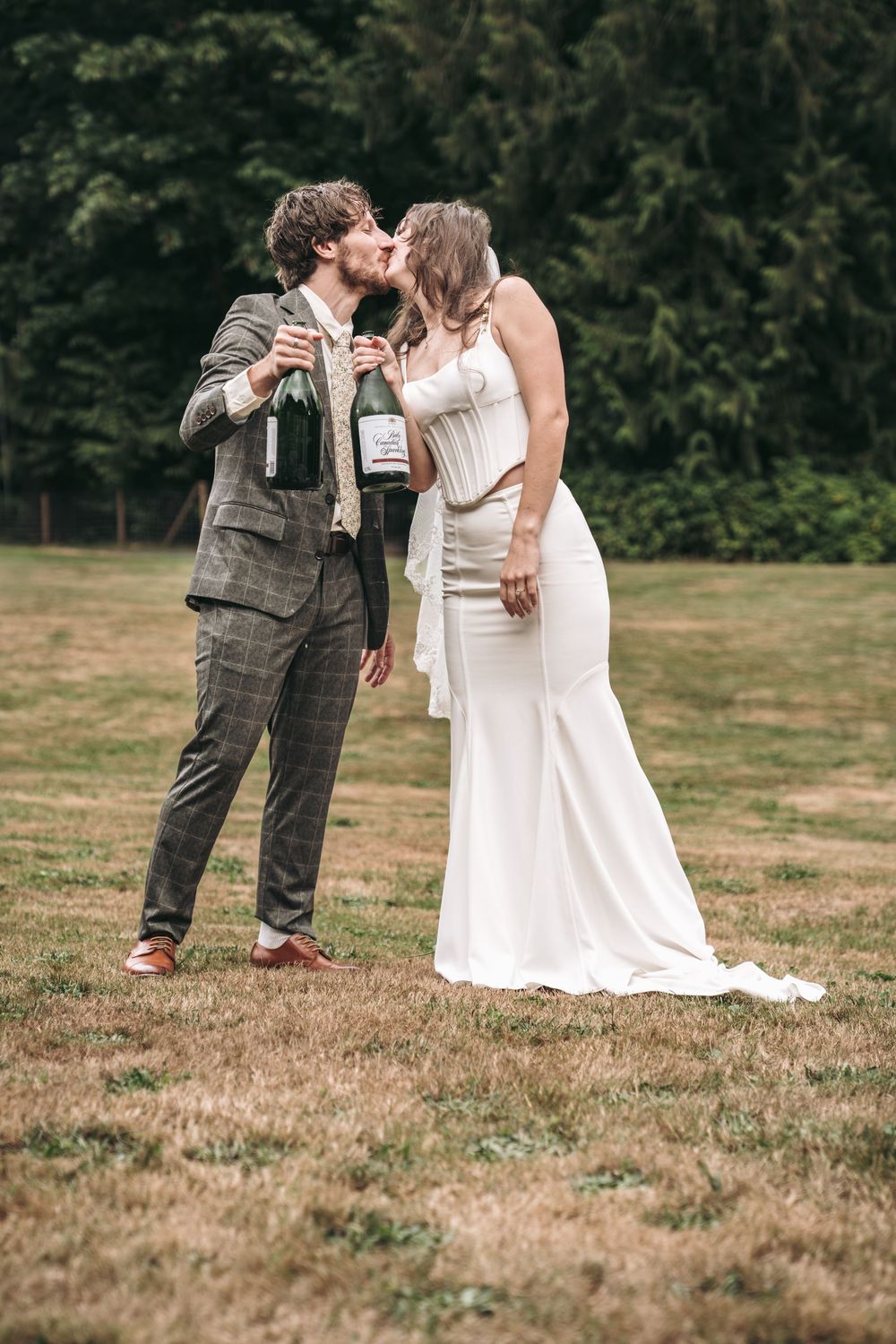 Bride and groom kissing outdoors on grass, holding champagne bottle, wearing white dress and plaid suit.