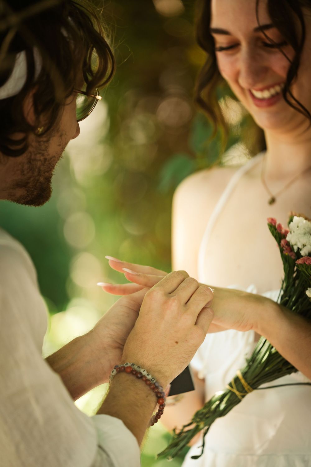 Groom placing wedding ring on smiling brides finger during outdoor ceremony in sunlit garden.
