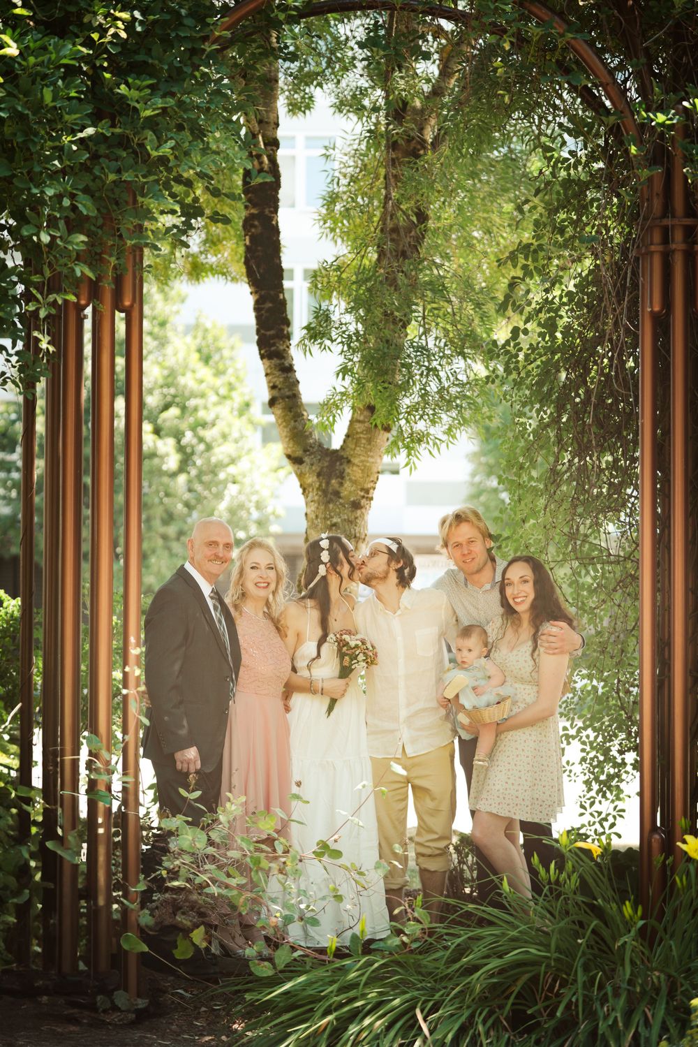 Wedding family portrait of six people posing under garden arbor surrounded by lush green foliage.