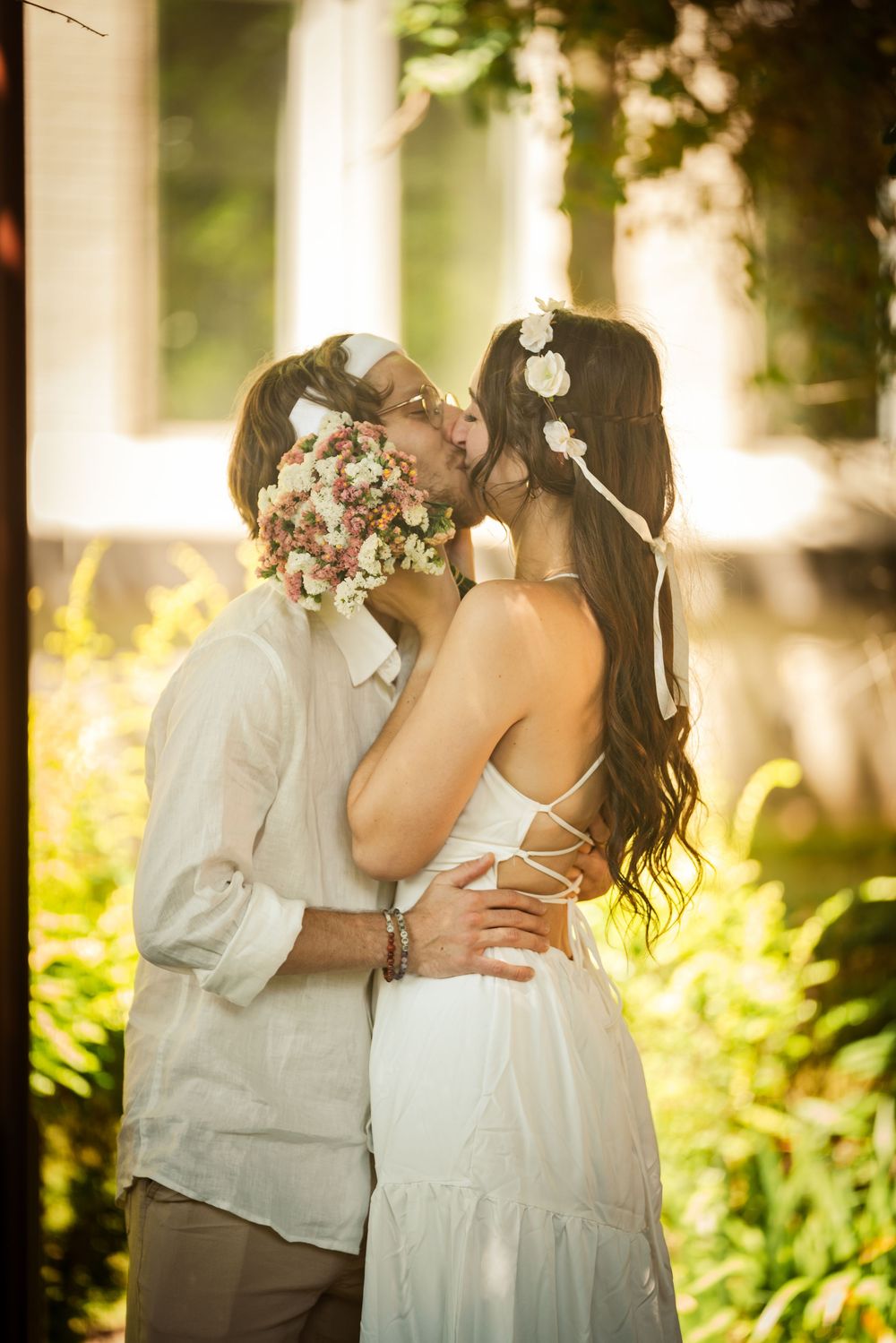 Newlywed couple sharing romantic kiss holding bouquet in sunlit garden, bride in white dress with floral crown.