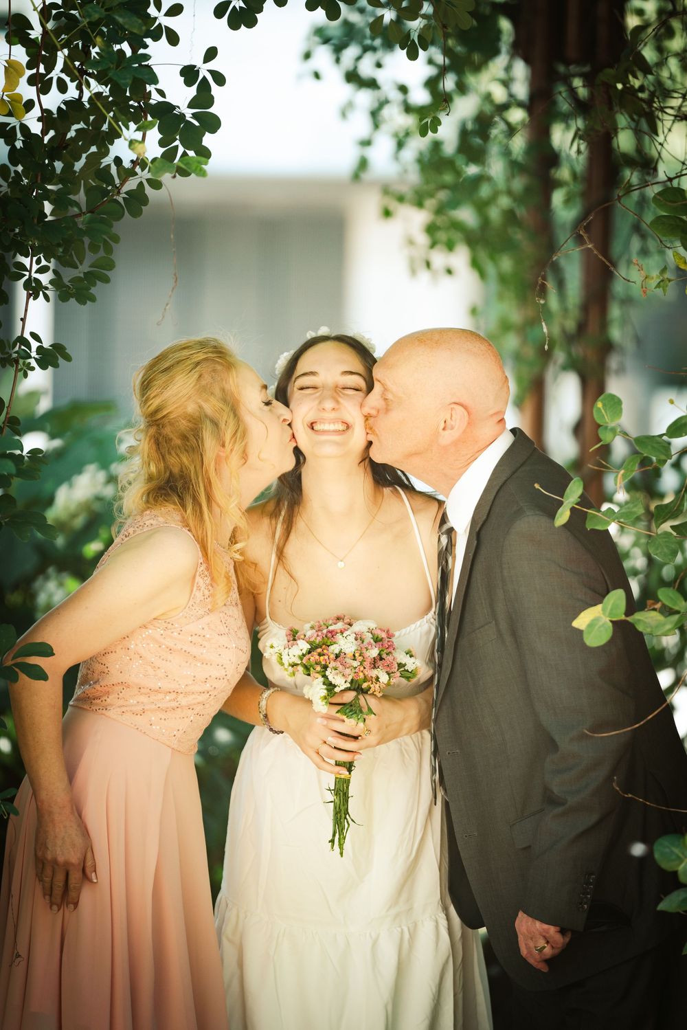 A smiling bride holding flowers is kissed on the cheeks by her parents under green leafy branches.