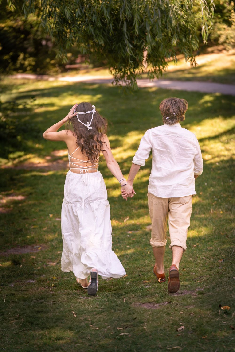 A bride and groom jog hand in hand away from the camera through a sunny park.