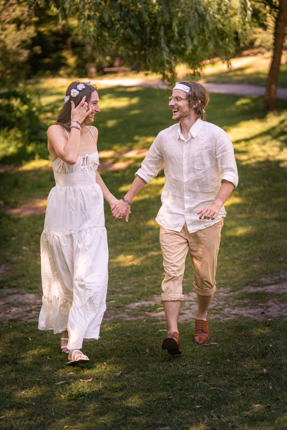 A bride and groom in bohemian attire holds hands and laughs while running through a lush green park.